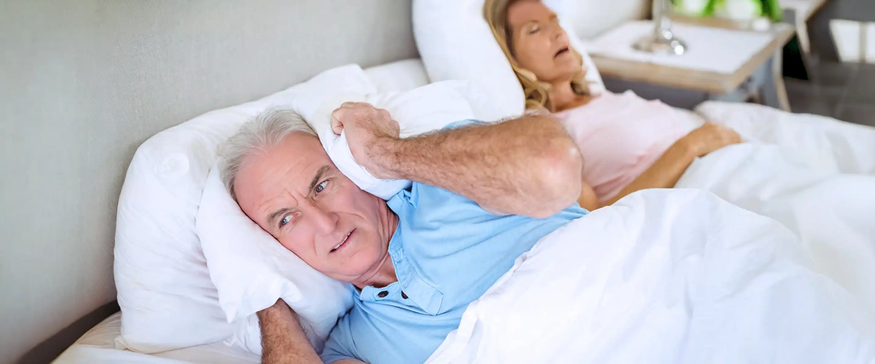 Senior man lying on bed and covering his ears with pillow in bedroom